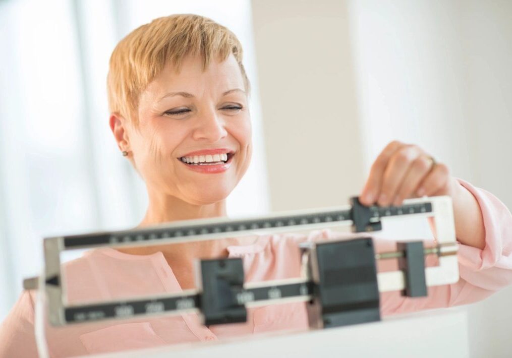 A woman is smiling while adjusting the weights on a balance scale in a brightly lit room.