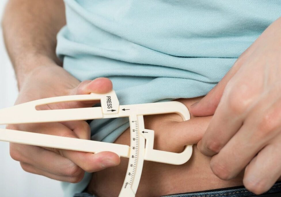 Person using a skinfold caliper to measure body fat percentage on the abdomen area, wearing a light blue shirt.